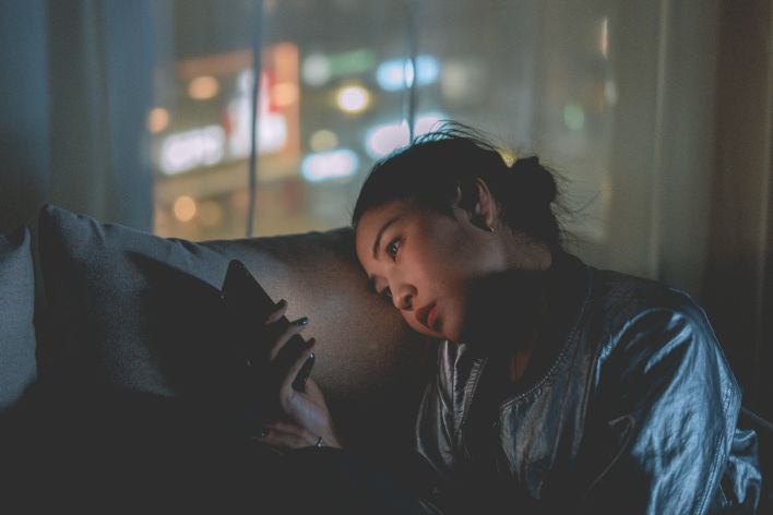 A woman looking at her mobile phone at night with city lights blurred in the background.