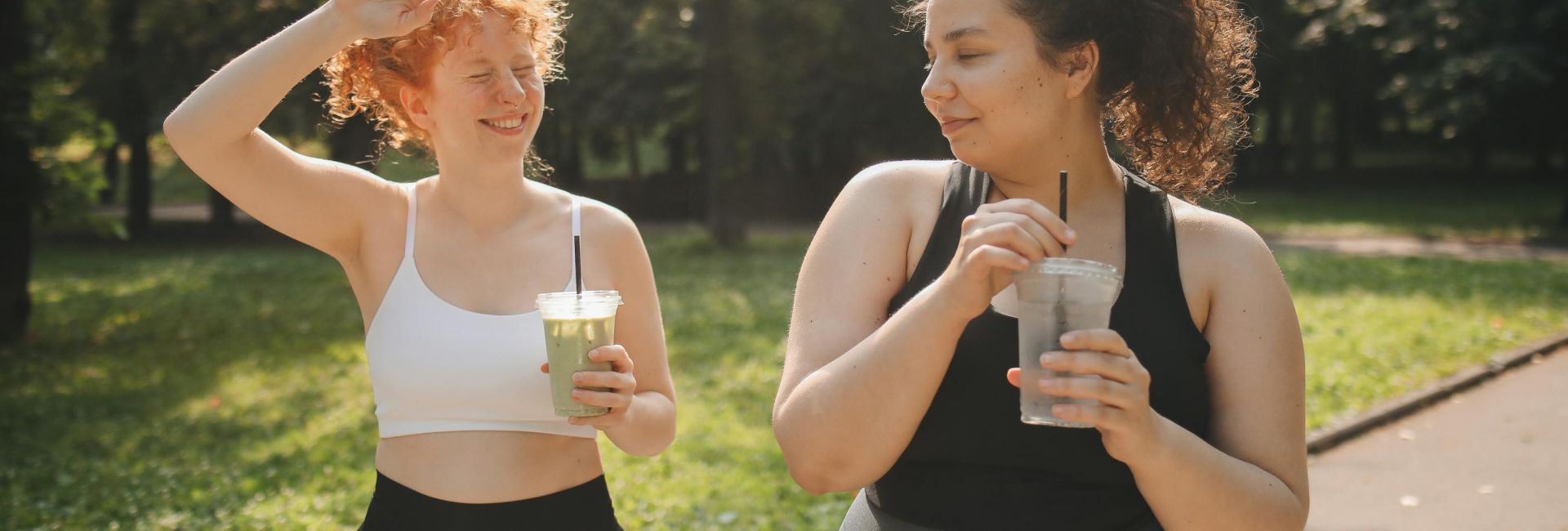Two women in activewear enjoy refreshing drinks while relaxing outdoors in a sunny park.