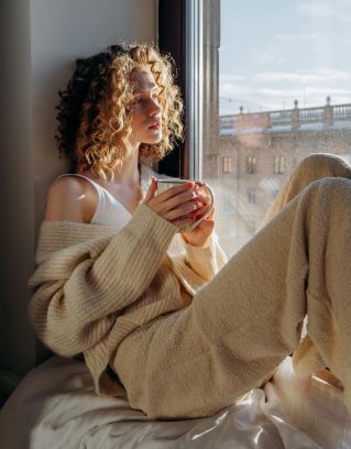 Woman in sweater sits by window, enjoying morning coffee with sunlight.