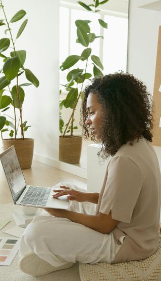 Curly-haired African American woman sitting with laptop, working in a modern indoor setting with plants.