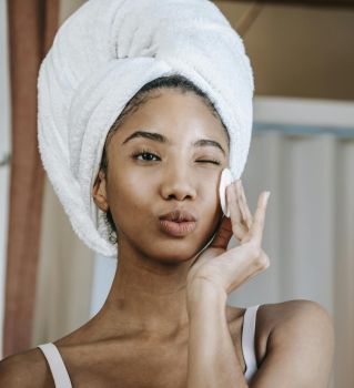 Charming ethnic female with folded lips winking while cleaning face with cotton pad during daily beauty routine
